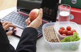 Una mano sostiene una manzana en un escritorio de trabajo, junto a un recipiente con verduras.