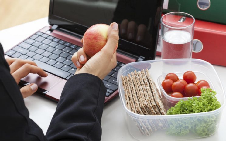 Una mano sostiene una manzana en un escritorio de trabajo, junto a un recipiente con verduras.