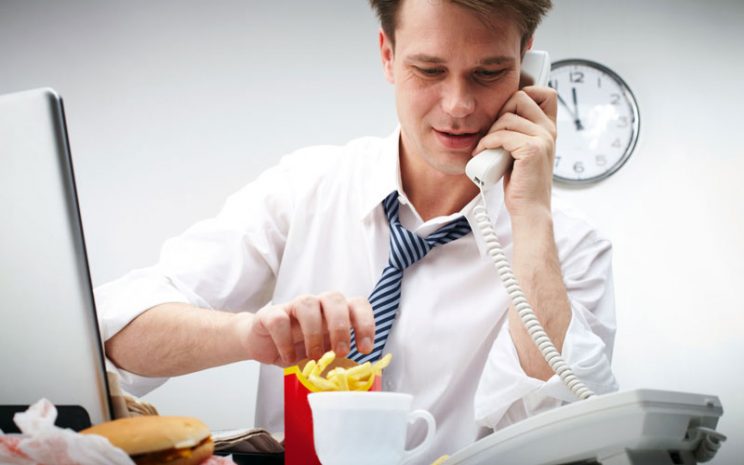 Oificinista come papas fritas sentado en su escritorio mientras habla por teléfono.