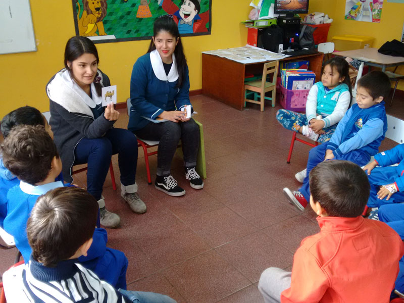 Dos estudiantes desarrollan un trabajo grupal con los niños sentados en círculo.