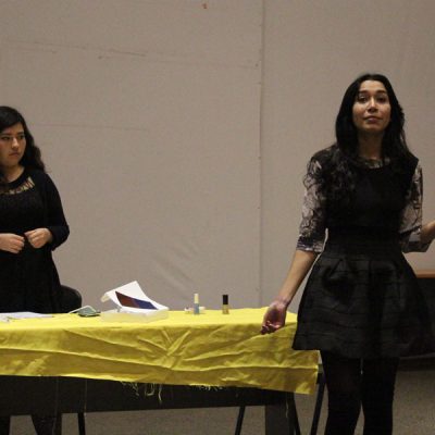 Una alumna habla y mueve su mano delante de una mesa decorada con un mantel amarillo. Atrás la observa una compañera.