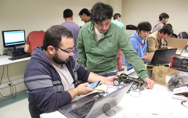 Dos estudiantes controlan un pequeño robot con ruedas mediante un teléfono celular.