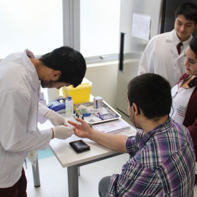 Tres estudiantes realizan análisis de grupo sanguíneo a alumno.