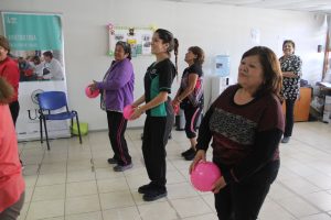 Dos adultas mayores realizan ejercicios con balón, y al medio, las acompaña una alumna de Kinesiología.