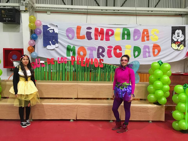 Dos alumnas de Santo Tomás, vestidas de hadas, posan frente un colorido cartel que dice Olimpiadas de Motricidad.