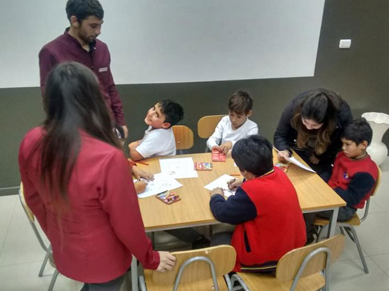 Tres niños dibujan sentados en una mesa, supervisados por tres alumnos de Psicología.