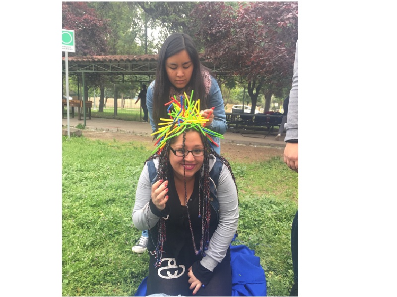 Una joven con lentes sonríe mientras su compañera le pone bombillas en el cabello, como parte de un juego.