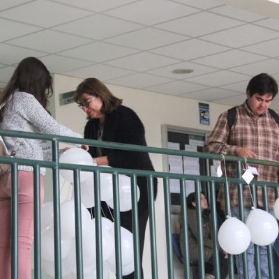 Estudiante instala globos en baranda, mientras dos personas preparan los globos.