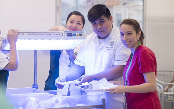 Una Matrona vestida de rojo, está junto a dos estudiantes de Técnico en Enfermería y una incubadora.