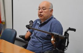Pablo Tapia, hombre canoso, con lentes, vestido de azul, habla al micrófono sentado en silla de ruedas eléctrica.