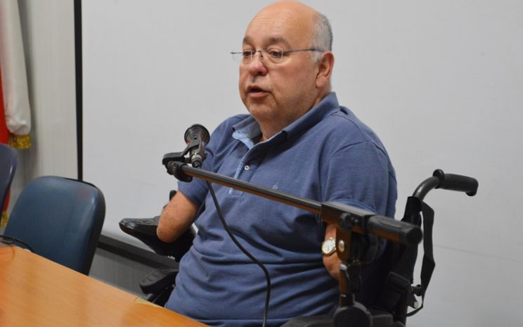 Pablo Tapia, hombre canoso, con lentes, vestido de azul, habla al micrófono sentado en silla de ruedas eléctrica.